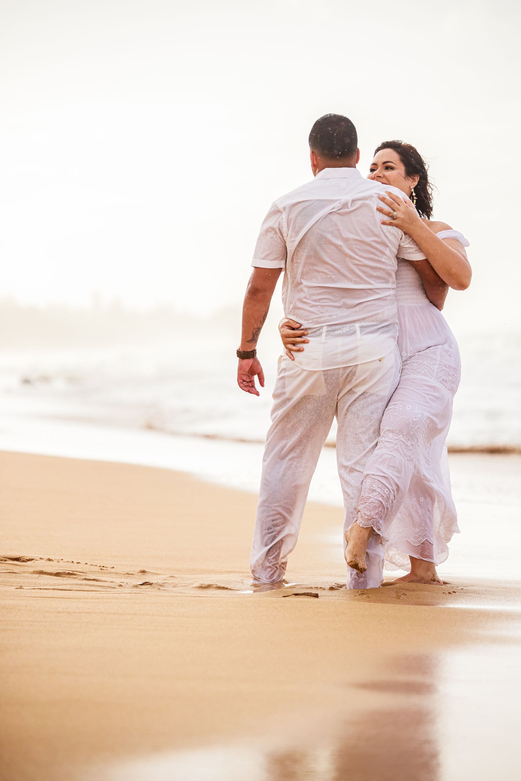 A couple in white clothes dances passionately by the ocean at sunset.